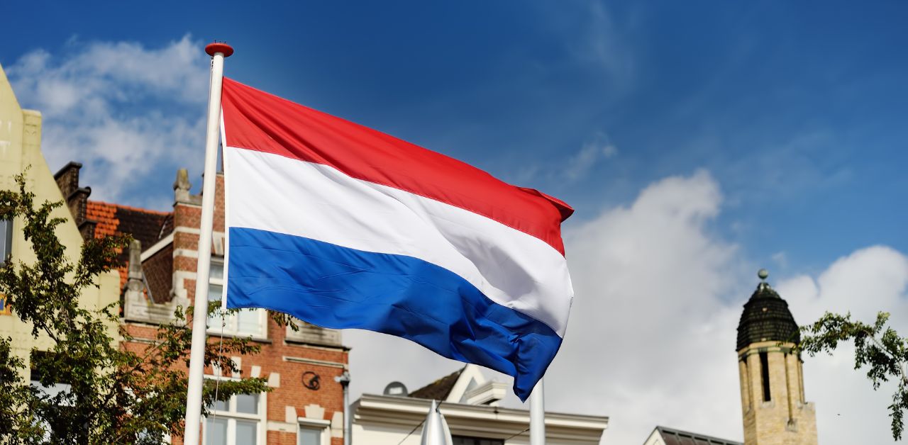 An image showing the flag of The Netherlands with a blue sky and buildings behind